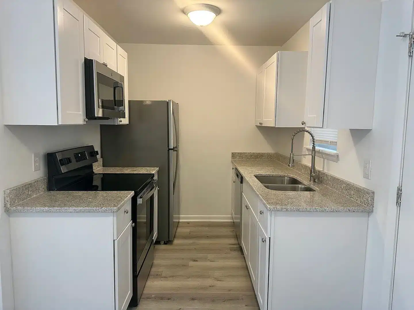 Modern galley kitchen with white cabinets, stainless steel appliances, granite countertops, and light wood flooring under ceiling light.