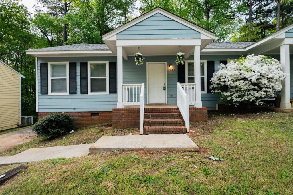 Light blue single-story house with white trim, small front porch with brick steps, hanging plants, and blooming white bush on the right, surrounded by trees and grass.