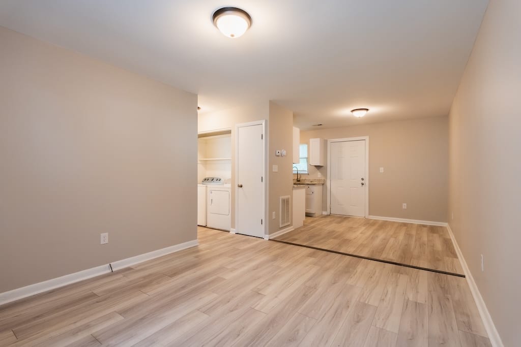 Unfurnished room with light wood flooring, beige walls, washer and dryer in a recessed area, and a white entry door near a small kitchen space.