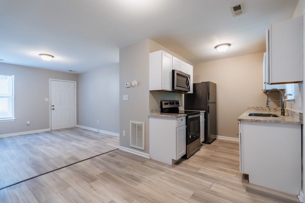 Modern kitchen with white cabinets, stainless steel appliances, and light wood flooring, open to a living area with a window and exterior door.
