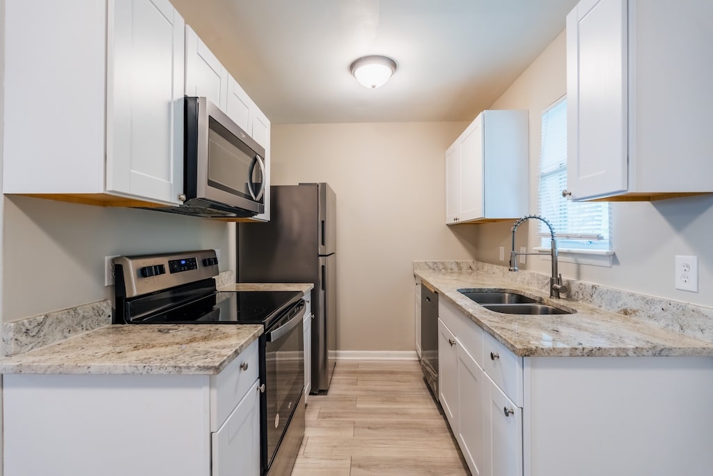 Modern kitchen with white cabinets, stainless steel appliances, electric stove, and a double sink under a window. Marble-patterned countertops and wood-look flooring are visible.