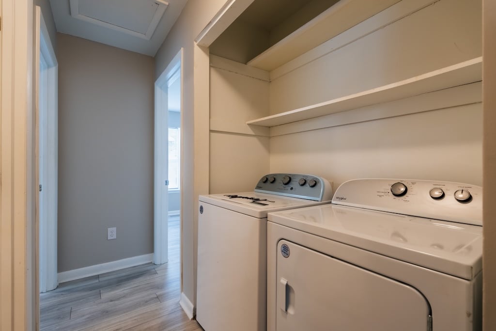 A laundry area with a washing machine and dryer beneath two built-in shelves is located in a hallway with neutral-colored walls and wood-style flooring.