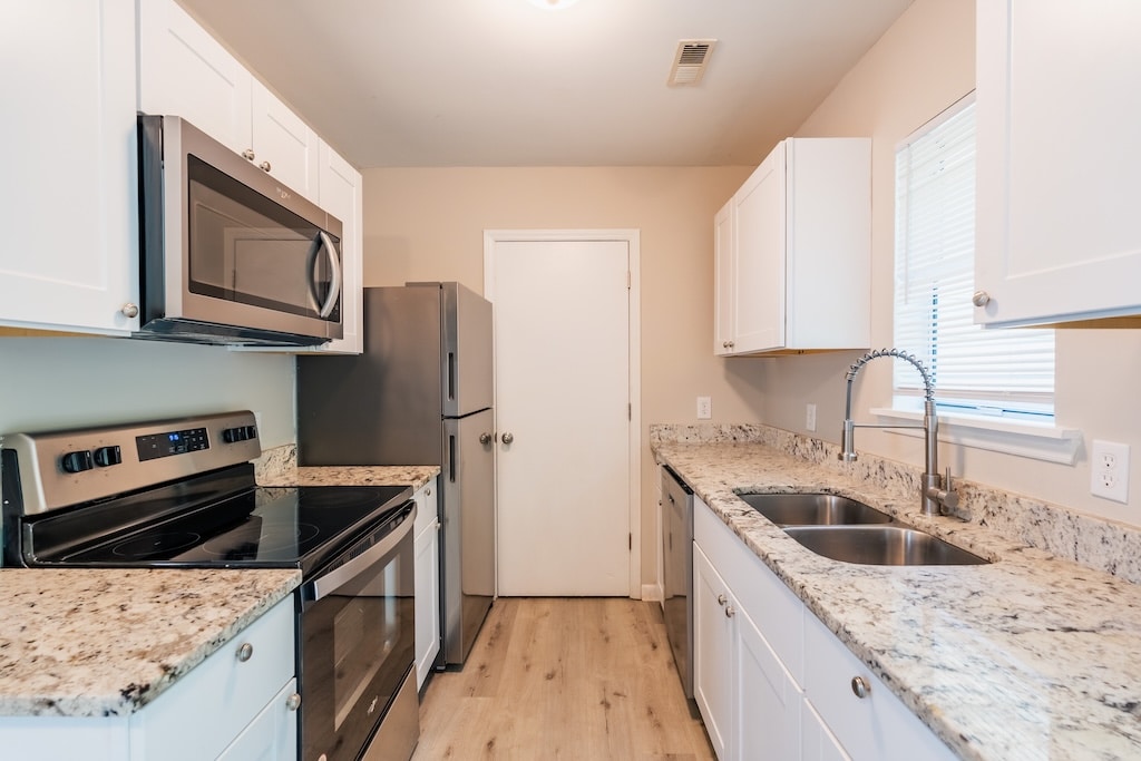 Modern kitchen with white cabinets, stainless steel appliances, granite countertops, double sink, and light wood flooring. Window above sink and a closed door at the end of the room.