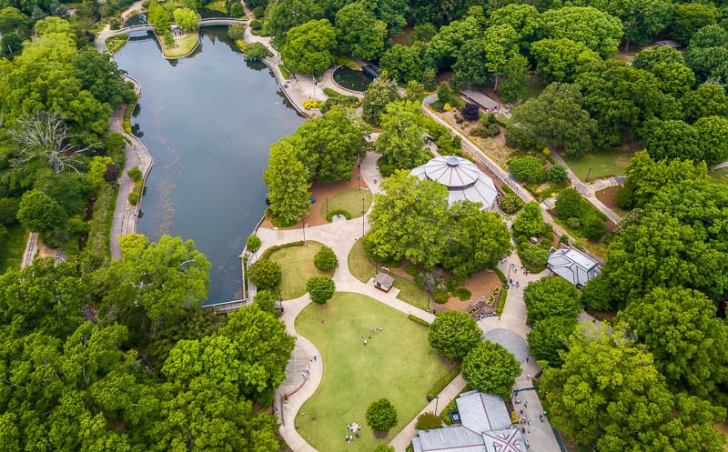 Aerial view of a green park with walking paths, a pond, open grassy areas, and several pavilion structures surrounded by trees.