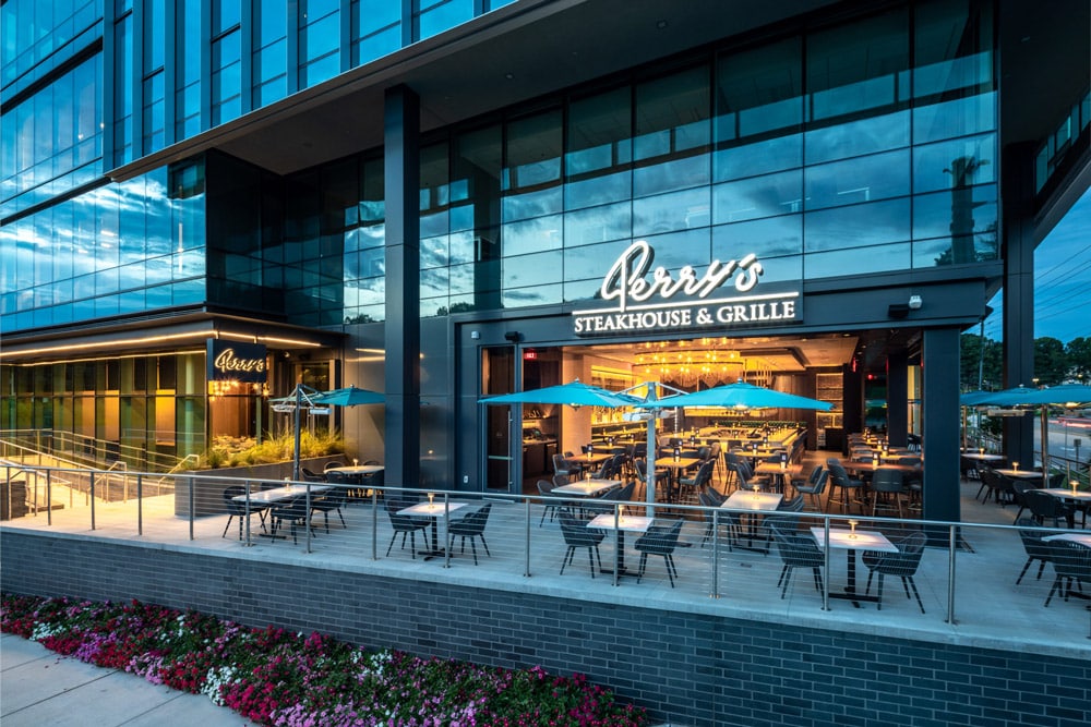 Outdoor patio seating and main entrance of Perry’s Steakhouse & Grille, a modern glass-front restaurant, with blue umbrellas and tables arranged on the terrace.